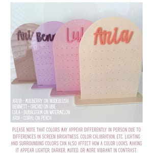 May include: Four earring display stands in pink and beige hues. Each stand features a name in a contrasting colour: Ariya, Bennett, Lula, and Aria. The stands have small holes for earrings and are on a rectangular base.
