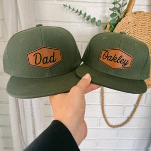 May include: Two green baseball caps with brown leather patches. The patch on the left cap says "Dad" and the patch on the right cap says "Oakley".