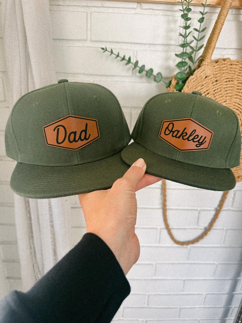 May include: Two green baseball caps with brown leather patches. The patch on the left cap says "Dad" and the patch on the right cap says "Oakley".