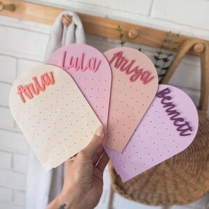 May include: Four pastel-coloured, arched earring display cards are held in a hand. Each card has a different name in a contrasting colour: Aria, Lyla, Ariya, and Bennett. The cards have small holes for earrings. A white brick wall is in the background.