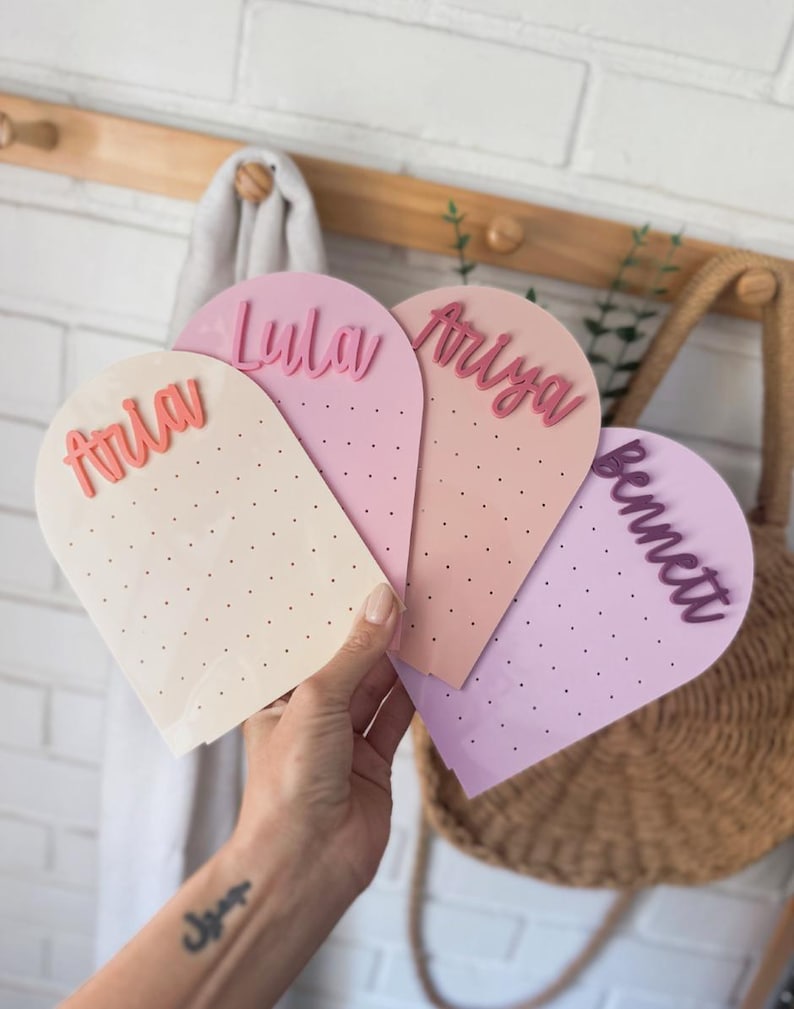 May include: Four pastel-coloured, arched earring display cards are held in a hand. Each card has a different name in a contrasting colour: Aria, Lyla, Ariya, and Bennett. The cards have small holes for earrings. A white brick wall is in the background.