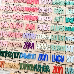 May include: A collection of wooden name plaques with colorful, raised lettering. Each plaque features a different name in a playful font, with the words "Look What Made" below. The names are in various colors, including pink, blue, and green.