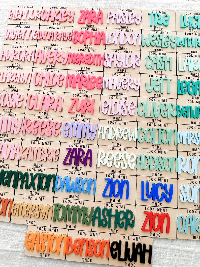 May include: A collection of wooden name plaques with colorful, raised lettering. Each plaque features a different name in a playful font, with the words "Look What Made" below. The names are in various colors, including pink, blue, and green.