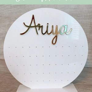 May include: White circular earring display stand with a gold mirror finish. The stand has a base and a round top with holes for earrings. The name "Ariya" is written in gold letters on the top of the stand.