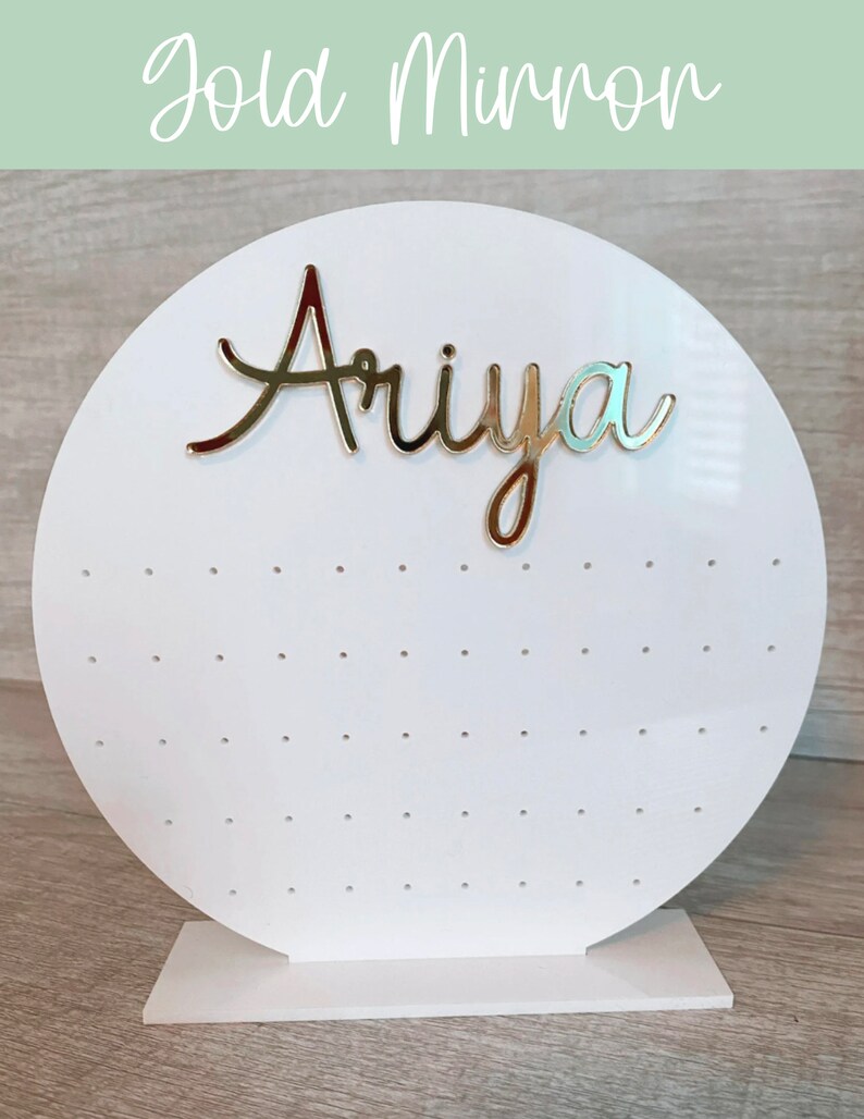 May include: White circular earring display stand with a gold mirror finish. The stand has a base and a round top with holes for earrings. The name "Ariya" is written in gold letters on the top of the stand.