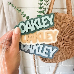 May include: Three name tags with the name "Oakley" in a bubble letter font. The tags are in shades of green, tan, and blue. The tags are held in front of a woven bag and a white brick wall.