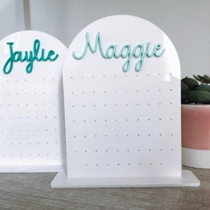 May include: Two white acrylic earring display stands with a rounded top. The stands have a grid of holes for holding earrings. The top of each stand has a name written in teal lettering. The stand on the left says "Jaylie" and the stand on the right says "Maggie".
