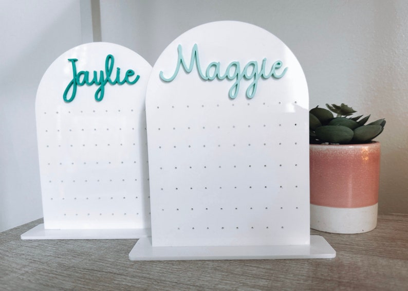 May include: Two white acrylic earring display stands with a rounded top. The stands have a grid of holes for holding earrings. The top of each stand has a name written in teal lettering. The stand on the left says "Jaylie" and the stand on the right says "Maggie".