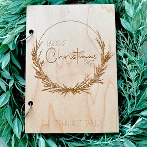 Pu&ograve; includere: Un album fotografico in legno con una rilegatura ad anelli. La copertina presenta un design inciso di una ghirlanda con il testo "Cards of Christmas Past" e "The Crowhurst Family".