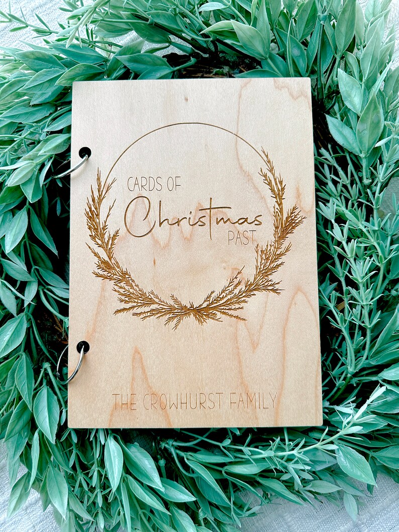 Pu&ograve; includere: Un album fotografico in legno con una rilegatura ad anelli. La copertina presenta un design inciso di una ghirlanda con il testo "Cards of Christmas Past" e "The Crowhurst Family".