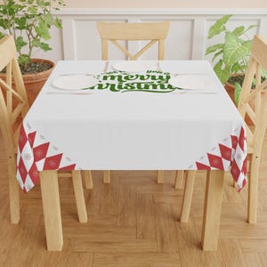 May include: A white tablecloth with a green "We wish you a Merry Christmas" text design. The tablecloth has a red and white diamond pattern with snowflakes on the corners. The table is set with plates and silverware.