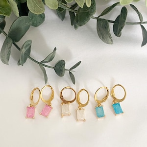 May include: Gold-coloured hoop earrings with rectangular, coloured gemstone pendants. The earrings are displayed in pairs, showcasing pink, white, and blue gemstones. Green foliage is in the background, set against a white backdrop.