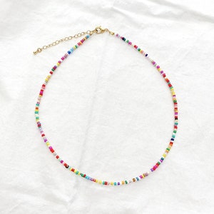 May include: A colorful beaded necklace with a gold-toned clasp and extender chain. The necklace features small, round beads in a variety of colors, including red, yellow, blue, green, and pink. The necklace is displayed on a white surface.