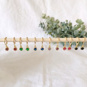 May include: A collection of gold-colored hoop earrings, each featuring a different colored gemstone. The gemstones include red, green, blue, and purple. The earrings are displayed on a wooden dowel against a white background.
