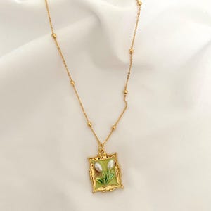 May include: A gold chain necklace with a gold frame pendant featuring a green and white enamel flower design. The pendant has two white pearl-like accents.