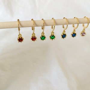 May include: A collection of gold-colored hoop earrings, each adorned with a different colored gemstone. The gemstones include red, green, and blue, and are suspended from the hoops. The earrings are displayed on a wooden dowel against a white background.