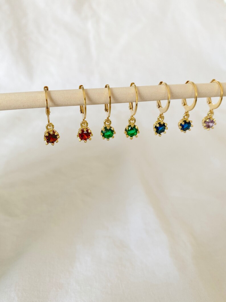 May include: A collection of gold-colored hoop earrings, each adorned with a different colored gemstone. The gemstones include red, green, and blue, and are suspended from the hoops. The earrings are displayed on a wooden dowel against a white background.