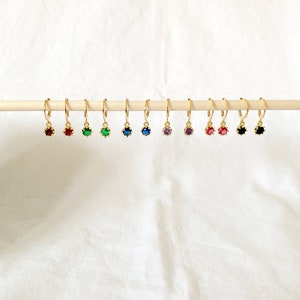 May include: A collection of gold-colored hoop earrings, each featuring a different colored gemstone. The gemstones include red, green, blue, purple, pink, and black. The earrings are displayed on a wooden dowel against a white background.