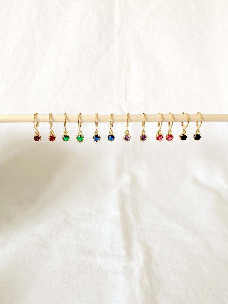 May include: A collection of gold-colored hoop earrings, each featuring a different colored gemstone. The gemstones include red, green, blue, purple, pink, and black. The earrings are displayed on a wooden dowel against a white background.