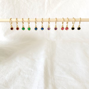 May include: A collection of gold-colored hoop earrings, each featuring a different colored gemstone. The gemstones include red, green, blue, purple, and black. The earrings are displayed on a wooden dowel against a white background.