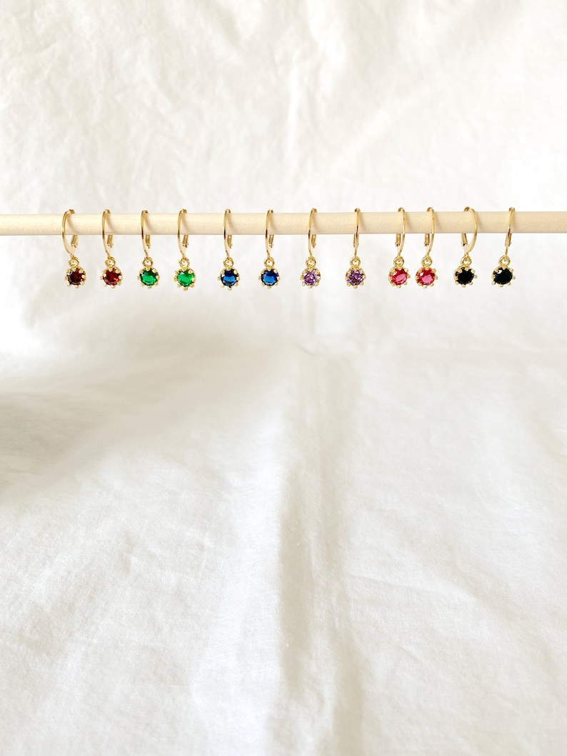 May include: A collection of gold-colored hoop earrings, each featuring a different colored gemstone. The gemstones include red, green, blue, purple, and black. The earrings are displayed on a wooden dowel against a white background.