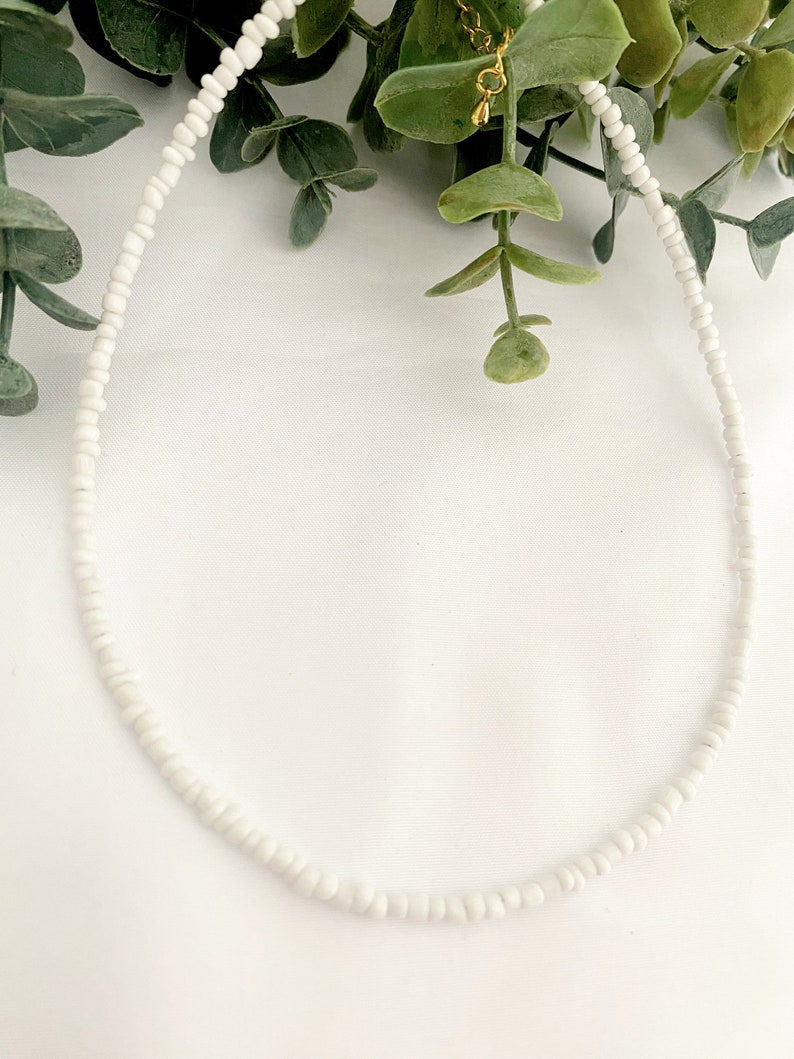 Peut inclure: Un d&eacute;licat collier de perles blanches. Le collier est compos&eacute; de petites perles blanches rondes enfil&eacute;es ensemble. Le fermoir et la cha&icirc;ne sont de couleur dor&eacute;e. Le collier est pr&eacute;sent&eacute; sur un fond blanc avec du feuillage vert.