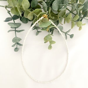 Peut inclure: Un d&eacute;licat collier de perles blanches avec un fermoir dor&eacute; et un petit charme. Le collier est pr&eacute;sent&eacute; sur un fond de feuillage vert et une surface blanche, cr&eacute;ant une esth&eacute;tique minimaliste.