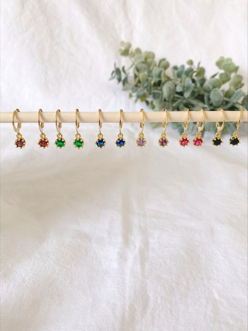 May include: A collection of gold-colored hoop earrings, each featuring a different colored gemstone. The gemstones include red, green, blue, and purple. The earrings are displayed on a wooden dowel against a white background.