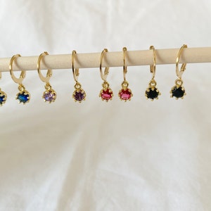 May include: Gold-colored hoop earrings with a small, colorful gemstone pendant. The gemstones are in various colors, including blue, purple, red, and black. The earrings are displayed on a wooden dowel against a white background.