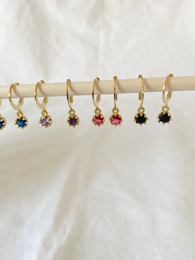 May include: Gold-colored hoop earrings with a small, colorful gemstone pendant. The gemstones are in various colors, including blue, purple, red, and black. The earrings are displayed on a wooden dowel against a white background.