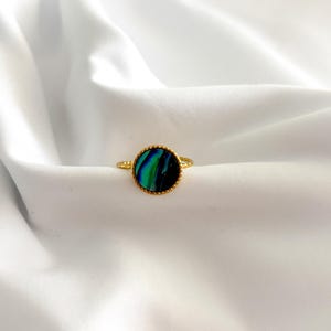 May include: A gold ring featuring a circular, iridescent stone in blues and greens. The stone is set in a gold bezel with a beaded border. The ring is presented on white, textured fabric.