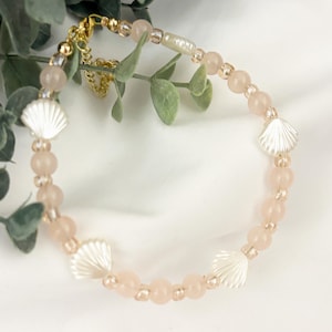 May include: A beaded bracelet featuring round, pale pink translucent beads interspersed with small clear spacer beads and four white, fan-shaped seashell charms. The bracelet includes a gold-toned metal lobster clasp and an adjustable chain link.