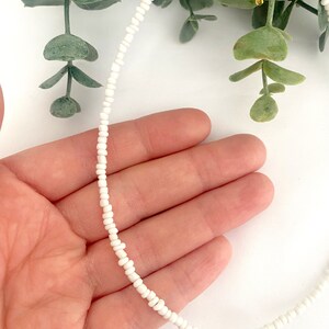Peut inclure: Un collier de perles blanches d&eacute;licat. Le collier est compos&eacute; de petites perles blanches espac&eacute;es uniform&eacute;ment. Le collier est tenu dans une main, avec un feuillage vert en arri&egrave;re-plan. Un accessoire simple et &eacute;l&eacute;gant.