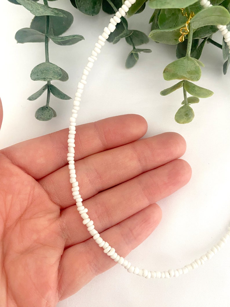 Peut inclure: Un collier de perles blanches d&eacute;licat. Le collier est compos&eacute; de petites perles blanches espac&eacute;es uniform&eacute;ment. Le collier est tenu dans une main, avec un feuillage vert en arri&egrave;re-plan. Un accessoire simple et &eacute;l&eacute;gant.