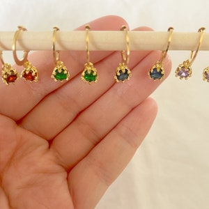 May include: A collection of gold-colored hoop earrings, each adorned with a different colored gemstone. The gemstones include red, green, blue, and purple. The earrings are displayed on a wooden dowel against a soft, neutral background.