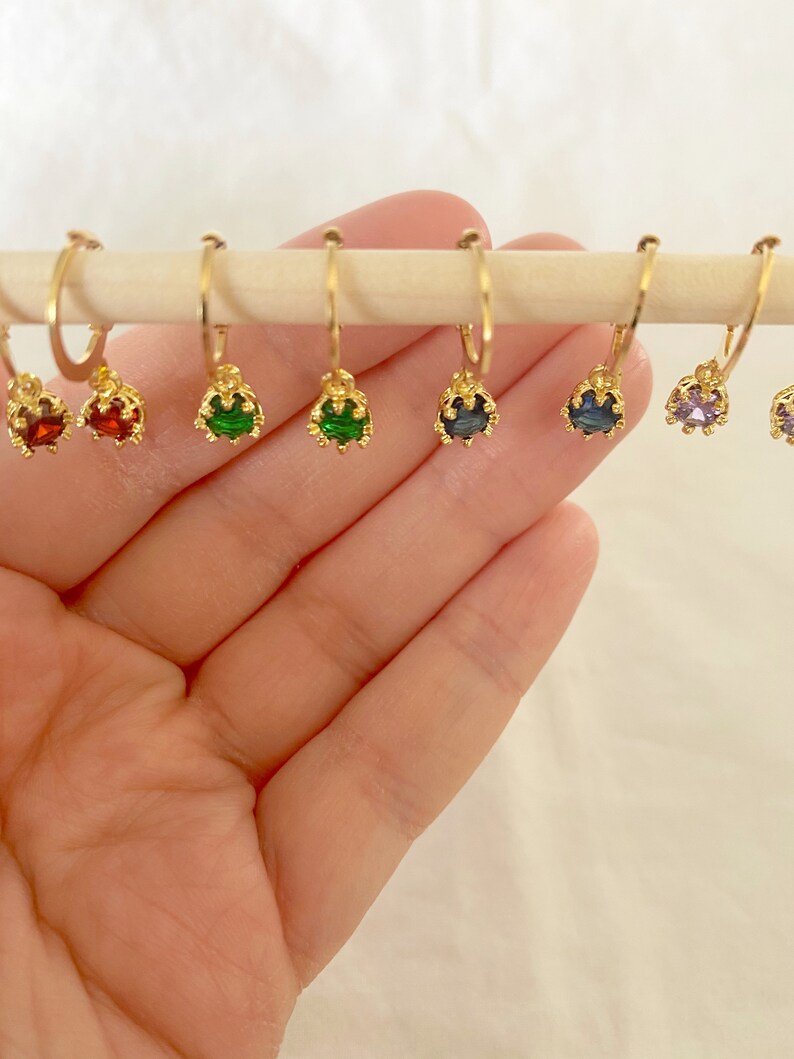 May include: A collection of gold-colored hoop earrings, each adorned with a different colored gemstone. The gemstones include red, green, blue, and purple. The earrings are displayed on a wooden dowel against a soft, neutral background.