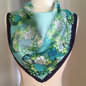 May include: A turquoise blue silk scarf with a floral pattern in shades of green, white, and blue. The scarf has a navy blue border.