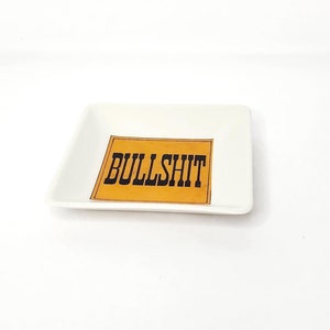 May include: A small, square, white ceramic dish with the word "BULLSHIT" printed in black block letters on a yellow rectangular background. The dish is centered on a white background.