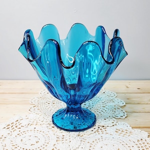 May include: A vibrant turquoise glass bowl with a ruffled rim and a pedestal base. The bowl sits on a white lace doily, displayed on a light wood surface. The bowl's dimensions are approximately 8 inches in diameter and 6 inches in height.