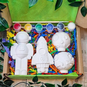 May include: A craft kit containing five white ceramic figurines: an astronaut, a rocket, a moon, and two planets. The kit also includes six paint pots with red, yellow, blue, green, and white paint, and a paintbrush.