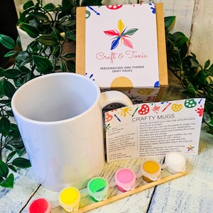 May include: A white ceramic mug, a wooden paintbrush, and six small pots of paint in red, yellow, green, pink, gold, and white. The set includes a cardboard box labelled Craft & Tonic and an instruction card titled Crafty Mugs with text.