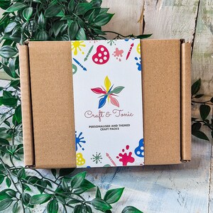 May include: A brown cardboard box with a colorful label that says "Craft & Tonic" and "Personalised and Themed Craft Packs". The label has a design of paintbrushes, paint palettes, and stars.