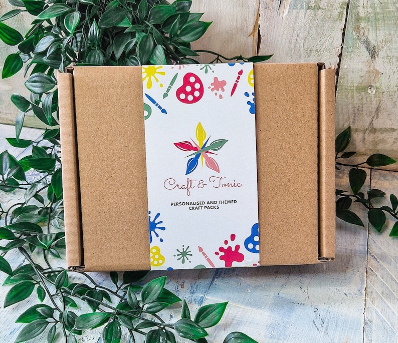 May include: A brown cardboard box with a colorful label that says "Craft & Tonic" and "Personalised and Themed Craft Packs". The label has a design of paintbrushes, paint palettes, and stars.