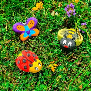 Puede incluir: Tres decoraciones de jard&iacute;n pintadas: una mariposa p&uacute;rpura y naranja, una mariquita roja y negra y una abeja amarilla y negra. Las decoraciones est&aacute;n sobre un lecho de hierba verde.