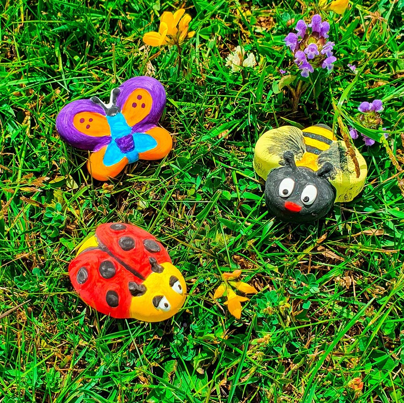 Puede incluir: Tres decoraciones de jard&iacute;n pintadas: una mariposa p&uacute;rpura y naranja, una mariquita roja y negra y una abeja amarilla y negra. Las decoraciones est&aacute;n sobre un lecho de hierba verde.