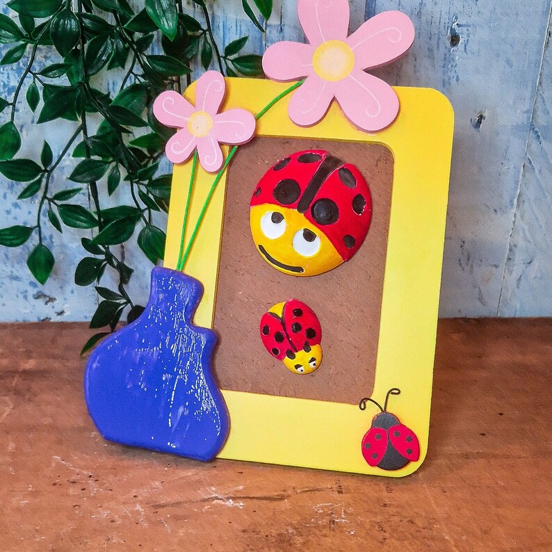 Puede incluir: Un marco de fotos de madera amarillo con un jarr&oacute;n azul y tres decoraciones de mariquitas. El marco tiene un fondo marr&oacute;n con un borde blanco. El jarr&oacute;n est&aacute; decorado con un patr&oacute;n floral blanco y azul. Las mariquitas son rojas con manchas negras y tienen ojos blancos.