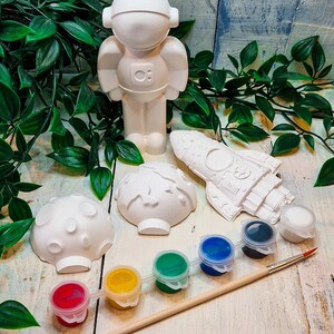 May include: A white ceramic astronaut figurine, rocket, and two planets, along with a paintbrush and six small paint pots in red, yellow, green, blue, and black.