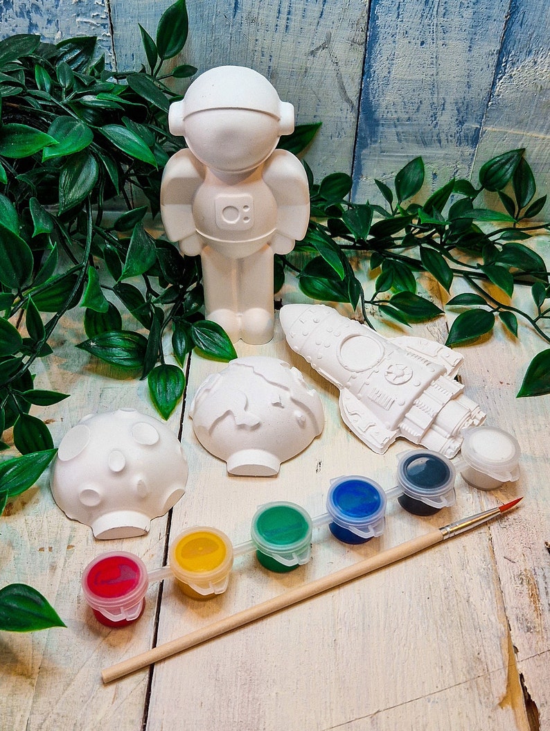 May include: A white ceramic astronaut figurine, rocket, and two planets, along with a paintbrush and six small paint pots in red, yellow, green, blue, and black.