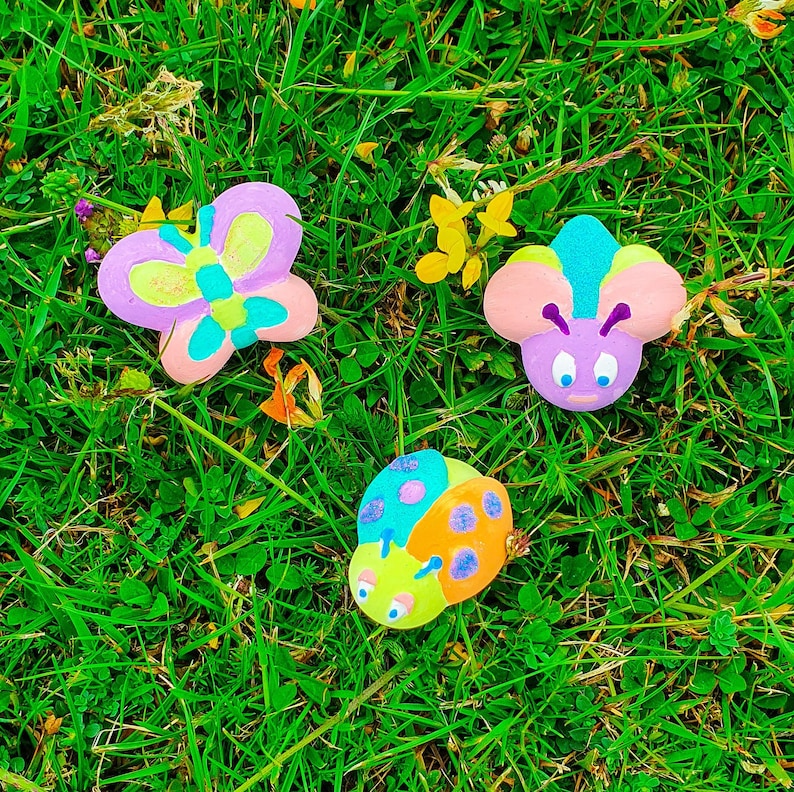 Puede incluir: Tres adornos de arcilla pintados de colores vivos en forma de mariposa, mariquita y abeja. Los adornos est&aacute;n sobre un lecho de hierba verde.