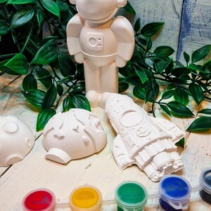 May include: A space-themed craft kit with unpainted figurines of an astronaut, a rocket ship, and a planet. The kit includes five small paint pots in red, yellow, green, blue, and black, and a paintbrush.